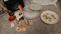 Lot items including one red Fiesta small bowl, three large Denby platters, hand-painted Shasta platter, assorted Chinese Yuan paper currency, New Hampshire toll coins, and some small boxes and tokens on carpeted floor.