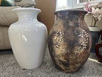 Two floor vases placed on carpet near window and couch. Shows white ceramic vase and brown composite vase with floral patterns.