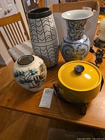 Four items on a wooden dining table: yellow electric fondue pot, white vase with Asian-style painting, tall white vase with black geometric pattern, blue and white ceramic vase.