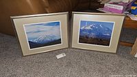 Two framed photographs of snowy mountain scenes, signed by Agranat, placed on carpeted floor.