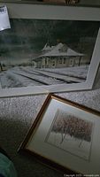 Photo showing two framed landscape artworks, one with a snowy train station and the other a snowy forest scene, placed on carpeted floor.