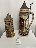 Photo showing two German beer steins, one taller with more ornate decoration and lid, one shorter with tavern scene.