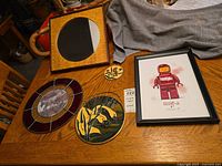 View showing all hanging decor items laid on a wooden table, including the wooden framed mirror, two stained glass pieces, small round plaque, and framed Lego print