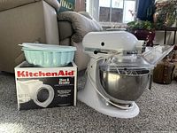 White KitchenAid K45SS stand mixer with metal bowl and clear slicer/shredder attachment next to a Tupperware Jell-O mold container on top of the KitchenAid slicer and shredder box.