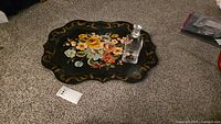Black metal tray on the floor showing hand-painted floral artwork and scalloped edges with the glass decanter on top.
