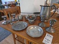 Full set of pewter items including 3 Steiff cups, plates, candlesticks, creamer, bowl, and pitcher on wooden table.