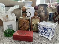 View of the full lot of items: Mikasa green glass paperweight, carved red trinket box, two vintage glass decanters, and blue-white ceramic planter on carpet.