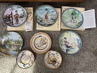 Overview of eight decorative plates on carpet showing oriental styled plates and scenic designs with boxes beneath some plates.