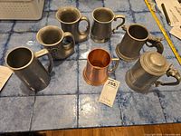 View showing all seven mugs on a blue tiled surface. Includes one copper mug and six pewter mugs with different styles; one pewter tankard has a hinged lid.