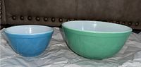 Two Pyrex mixing bowls, one smaller and blue with white interior, the other larger and green with white interior, placed side by side on crumpled paper.