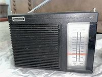 Front view of black Grundig FM/AM radio with speaker grill, tuning dial with frequency markings for AM and FM, and telescoping antenna extended.