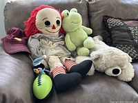 Photo showing Raggedy Ann doll seated on a couch with green frog plush, white teddy bear plush, small penguin stuffed toy, burgundy baseball cap, and green and black squishy football toy.