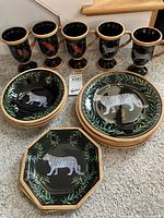 Five black glazed goblets with goldish brown rims, decorated with colorful safari animals including a leopard and a parrot, arranged in a row behind plates and bowls.