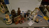 Wide view of assortment of religious figurines and decor on surface with a white cooler in background, includes Madonna and Child, angel, nativity box, and various small statuettes.