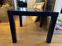 Front and side angle view of black square side table on wood floor, showing four legs and table top height.