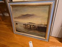 Framed oil painting leaning against wall on floor showing pier and water scene.