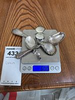 Several sterling silver spoons, a ladle, and a pill box on digital scale showing 60 grams.