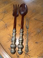 Photo of two large serving utensils with wood and silver plated handles alongside small silver plated fork on wooden table surface.
