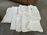 Group of folded white table linens including plain and patterned pieces laid on carpet.