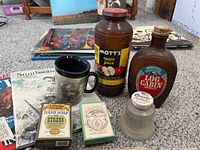 Photo showing various commemorative items including Mott's apple juice jar, Log Cabin syrup bottle, Liberty cherries jar, acrylic coffee cup, hand soap box, and green box with labeled design.