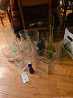 Wide shot of fourteen glass vases of various sizes and colors arranged on wooden floor.