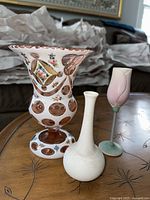 Three decorative vases displayed on a wooden table: a large pottery vase with floral and copper accents, a small white pottery bud vase, and a pink frosted glass bud vase with a green base.