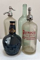 Five vintage glass bottles shown together: two soda siphons with metal Soda Stream tops, a clear glass bottle with red 'Pureoxia' text, a plain clear bottle with slight tint, and a dark blue embossed Chivas bottle made by Wade of England.