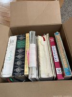 View of the top edges of seven vintage cookbooks and a folded fabric apron inside a cardboard box, showing their condition and arrangement.
