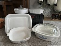 Photo showing three vintage CorningWare casserole dishes of white glasswear with blue floral design. One has glass lid, one shows visible chip.