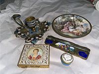 Photo showing the enamel metal candle holder with blue and white floral enamel on a leaf-shaped base, a rectangular enamel trinket dish with classical romantic imagery, a metal makeup compact with a painted female portrait on the lid, and a small round trinket dish inscribed 'Mum'.