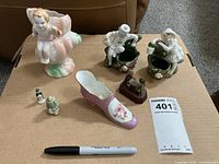Photo showing a collection of seven vintage ceramic figurines and a small mechanical decorative item arranged on a cardboard surface.