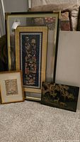 Full view of four items leaning on a couch: a tall framed embroidered panel, a gold-framed small print, a black panel with golden dragon, and a large blank frame.