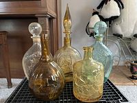 Five vintage glass decanters shown together on a grid surface, highlighting variety in shapes, colors, and stopper presence.