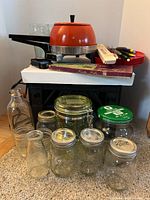 Vintage Hoover electric fry pan with orange enamel pot and lid, stacked on boxes with various kitchen items below and around it including glass bottles and jars.