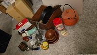 Overall lot showing wooden box, cast iron pan, orange cauldron, wooden bowls, tins, meat grinder, and smaller kitchen tools spread on floor.