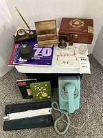 Photo showing various vintage office items on a table including a rotary phone, cigar box, notebooks, glasses, pen holder, bell and ink well.