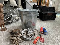 Lot overall with pulley, large metal pail, two flashlights, small garden leveler, and red garden trowel on carpeted floor