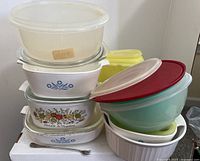 Stack of vintage glass Corning Ware and Pyrex dishes with some clear and red lids, plus yellow Tupperware canisters behind, and a stainless steel pickle fork in front