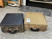 Two hard typewriter cases on a carpeted floor, one black and one beige, showing wear and dusty surfaces.
