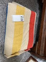 Folded cream-colored wool blanket with yellow, red, and blue stripes on carpeted floor.