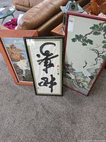 Three framed Asian art pieces leaning against a couch; two brown frames and one black frame are visible.