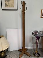 Full view of the wooden coat tree standing on a carpeted floor, showing the square vertical post and joined wooden base.