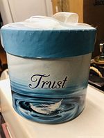 Photo of round box with blue water and feather design labeled 'Trust' and white ribbon bow on the lid.
