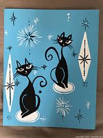 Full view of the 10x12 canvas painting showing two black stylized cats with white and black atomic starburst shapes on blue background.
