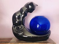 Side view of glossy black ceramic cat figurine sitting and curving its tail to hold a blue glass fishbowl.