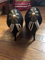 Back view of black cloth stilettos showing gold-tone flower with central pearl and hanging pearl strands detail on heel backs.