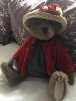 Full body shot of the Boyd's Lady Bear stuffed animal sitting upright on a gray floral blanket, showing the red cardigan, blue polka dot dress, and straw hat with flowers.