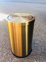 Front view of the cylindrical brass-colored metal garbage can showing its brushed finish and round flip top lid.