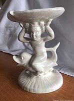 Off-white pearl iridescent ceramic soap dish featuring a cherub riding a dolphin, viewed from the front left side showing cherub's detailed facial features and dolphin's head.