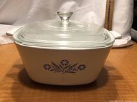 Square white Corningware casserole dish with Cornflower Blue floral pattern on the side and clear glass lid on top, shown on a surface.